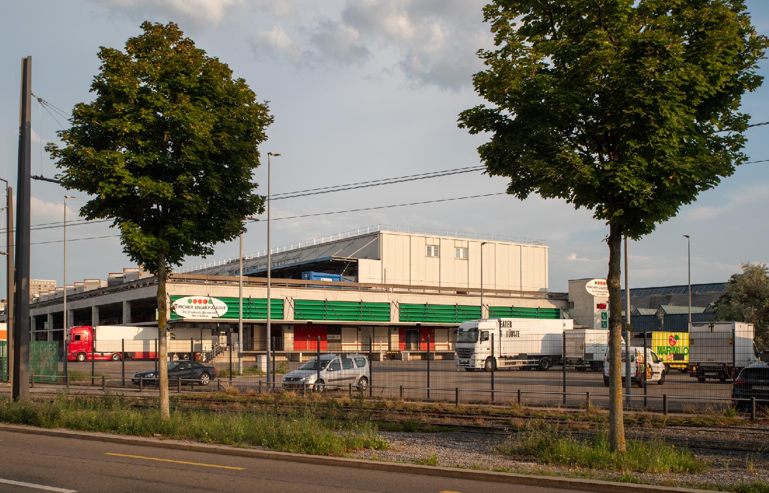 Docking bay of the Zürcher Three-story building, with a bunch of truck containers on the last floor. The building has flashy green blinds on the second floor. There is a road and a couple of trees in the foreground.