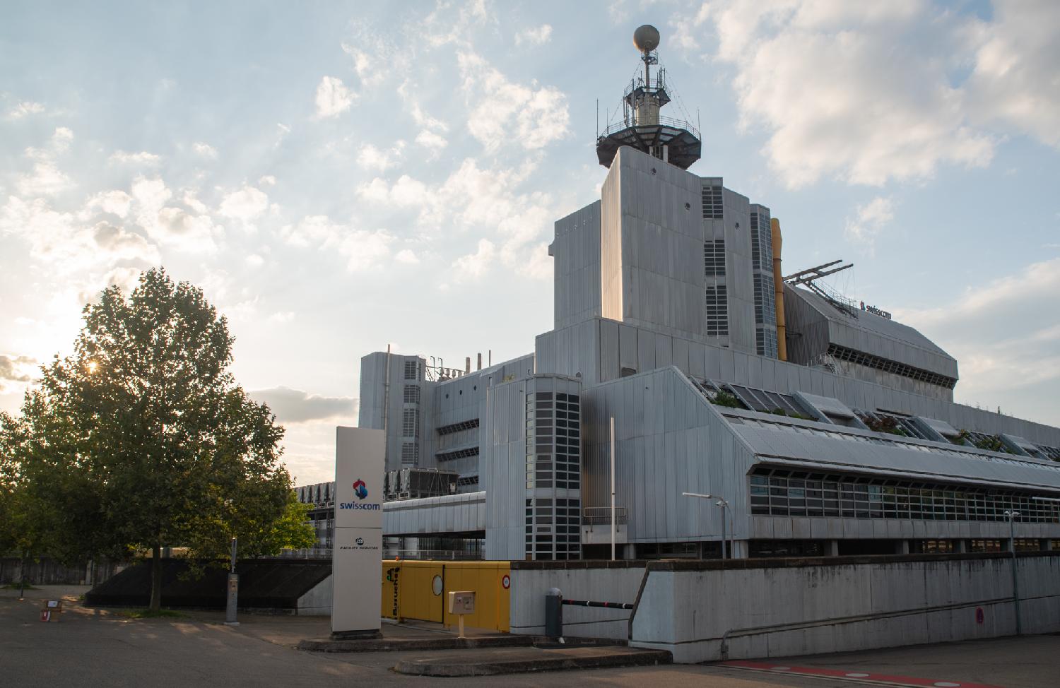 Swisscom datacenter, a large industrial building, made out of a bunch of blocks, with a large antenna-looking structure on top. The sun is quite low and visible through the leaves of the tree on the left of the building.
