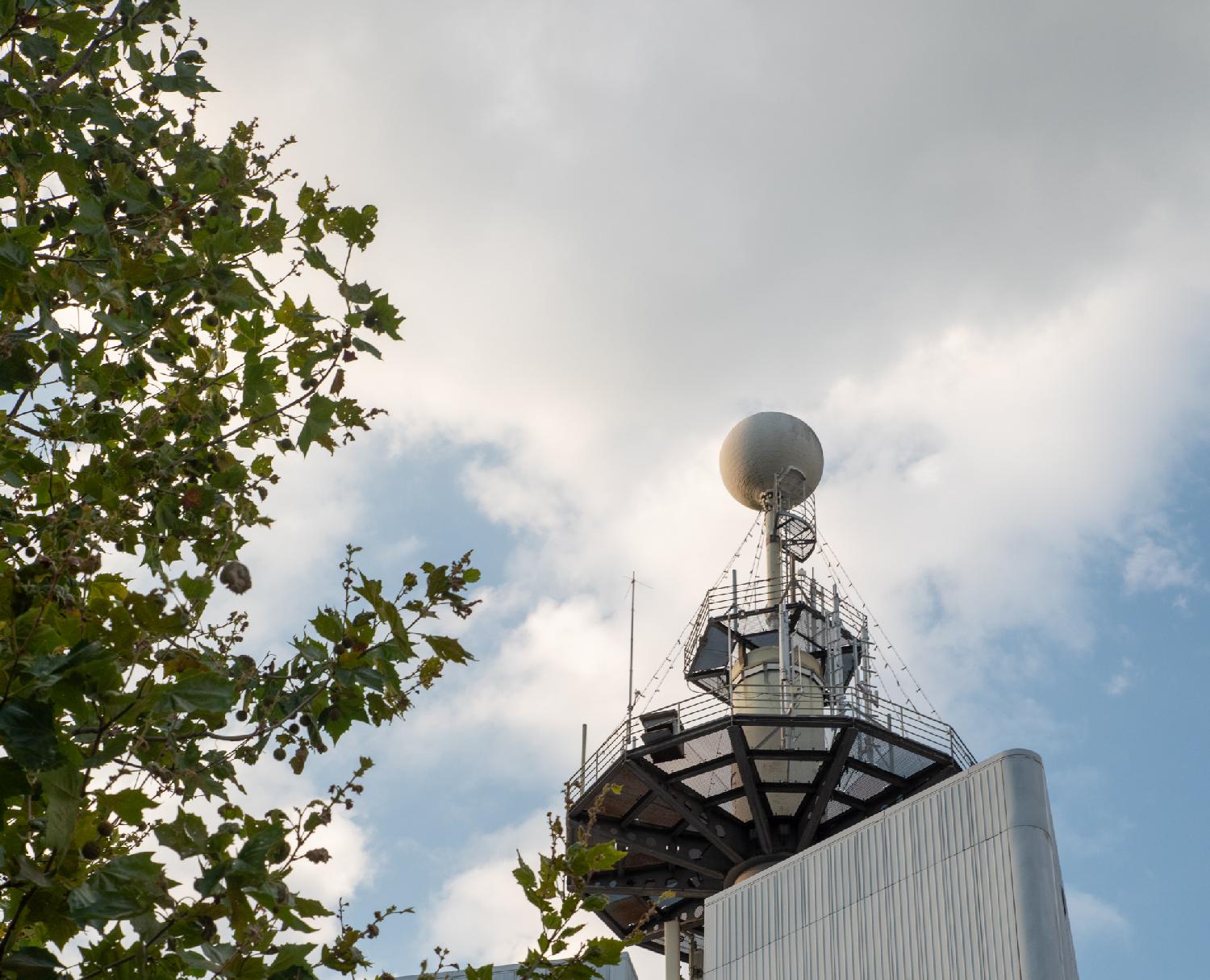 Top of the Swisscom building. A ball structure is on top of pole, with a small hexagonal platform below it, and a larger polygonal structure below it. The image is framed on the left by the leaves of a neighboring tree.