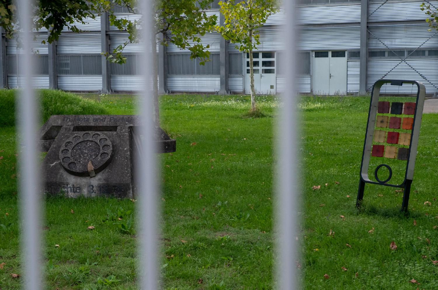 Two sculptures on a lawn: a large stone rotary phone, and a transparent smartphone with rows of colorful icons. The picture is taken through metallic grating. The building is visible in the background.