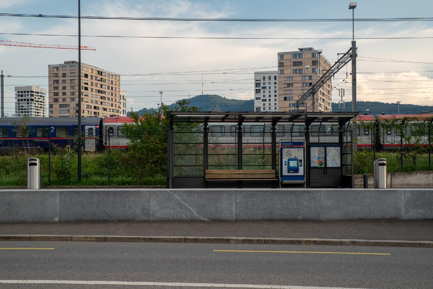 Empty tram stop, with a glass shelter, a wooden bench, and a ticket vending machine. The background has, in order, a passenger train, four high-rise buildings, and the Uetliberg.