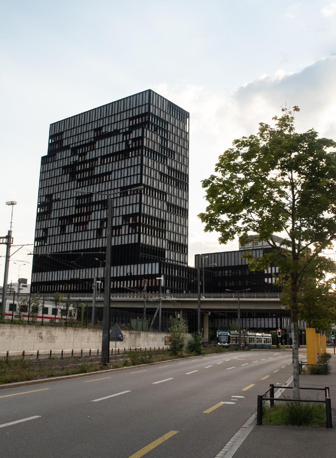 A small section of Aargauerstrasse with a ~20-story bulding, a bridge above the street, and a tram traveling on one side of the street.