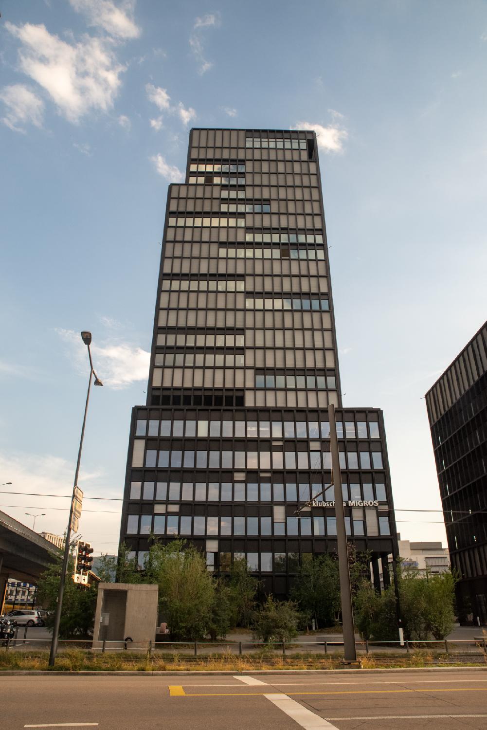 A 20+-story building, composed of a larger 5-story-base and a few blocks on top of it. Most windows have blinds in front of them; the others reflect the blue sky.