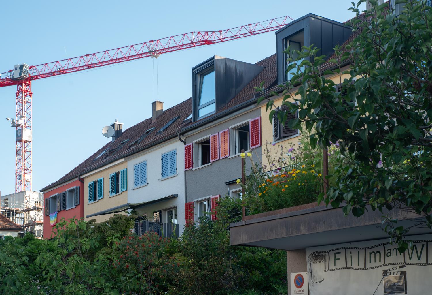 5 houses behind greenery, painted in red/orange, light yellow, white, grey and yellow. A rainbow peace flag hangs from one of the windows. There's a red crane in the background.