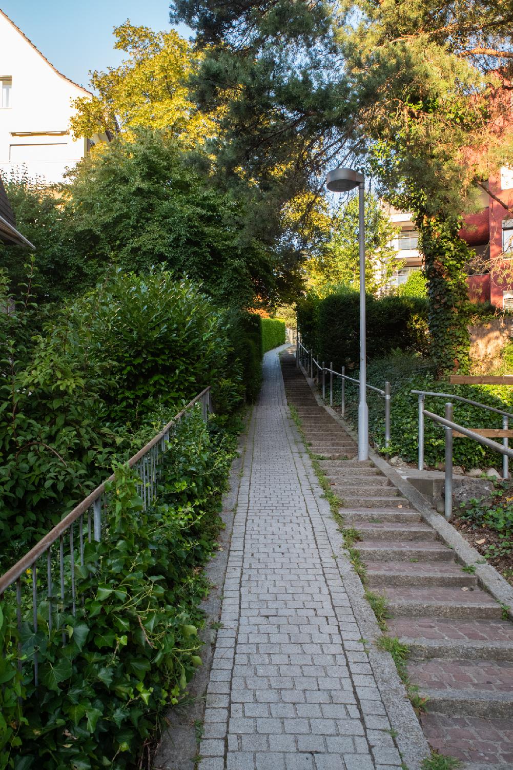 A small pedestrian alley, with grey blocks on the left half and dark red block steps on the right. The path goes up an turns slightly right at the end of the path in the middle of the picture. There are trees and greenery all around, and a few habitation buildings on each side.