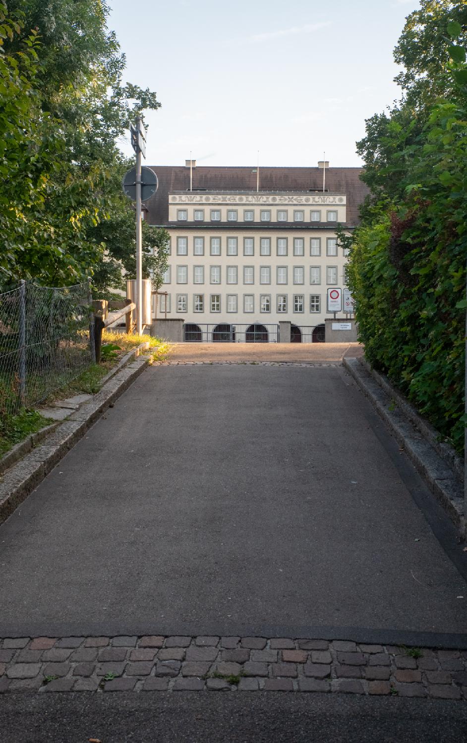 An imposing off-white building with decorative moldings above the top windows is visible between two rows of trees.