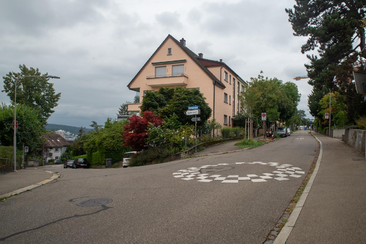 Intersection between two roads at an acute angle. The intersection is marked by a black and white circle on the road. A three-storey light-orange house building is on the corner of the intersection.