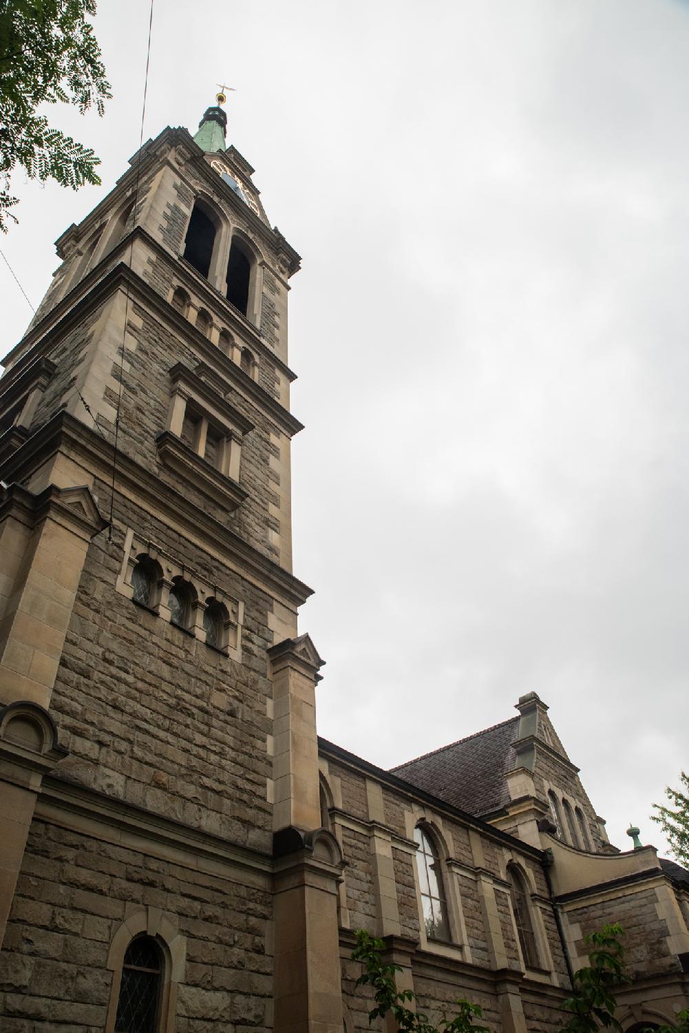 Church tower and building, viewed from the side. The church is built with beige stone and has windows with round tops. The tip of the church tower is barely visible due to the perspective and is made of green copper.