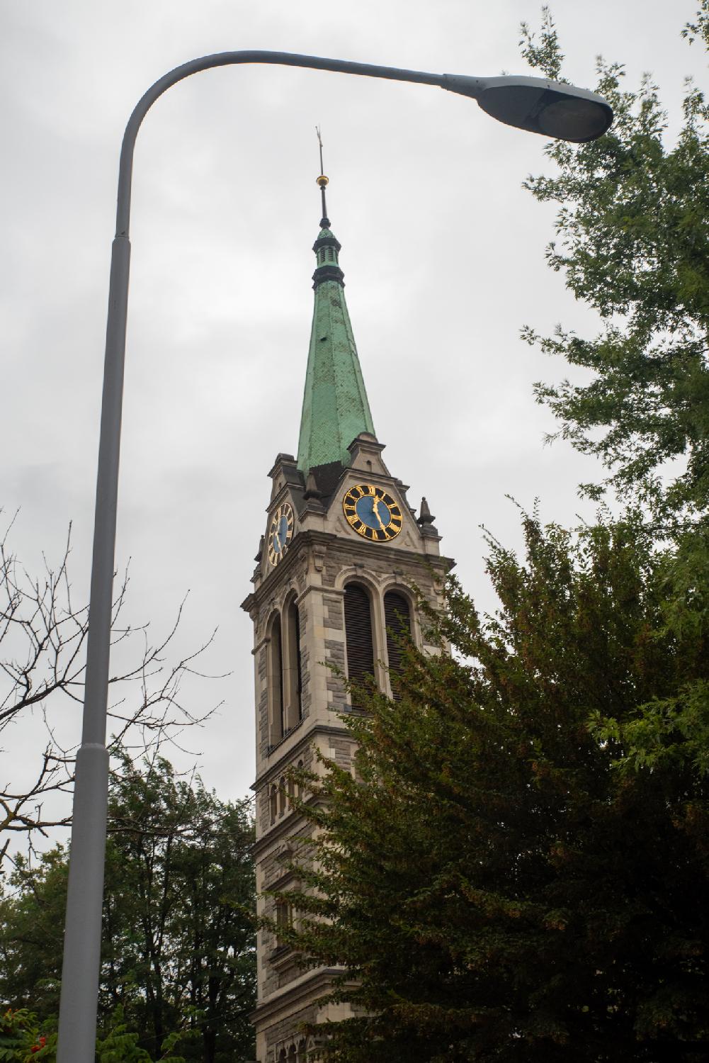 Church tower with a copper green spire, a blue and gold clock on each side showing half past twelve, and round-topped windows. The church tower is framed by a curved street lamp on the left and trees on the right.