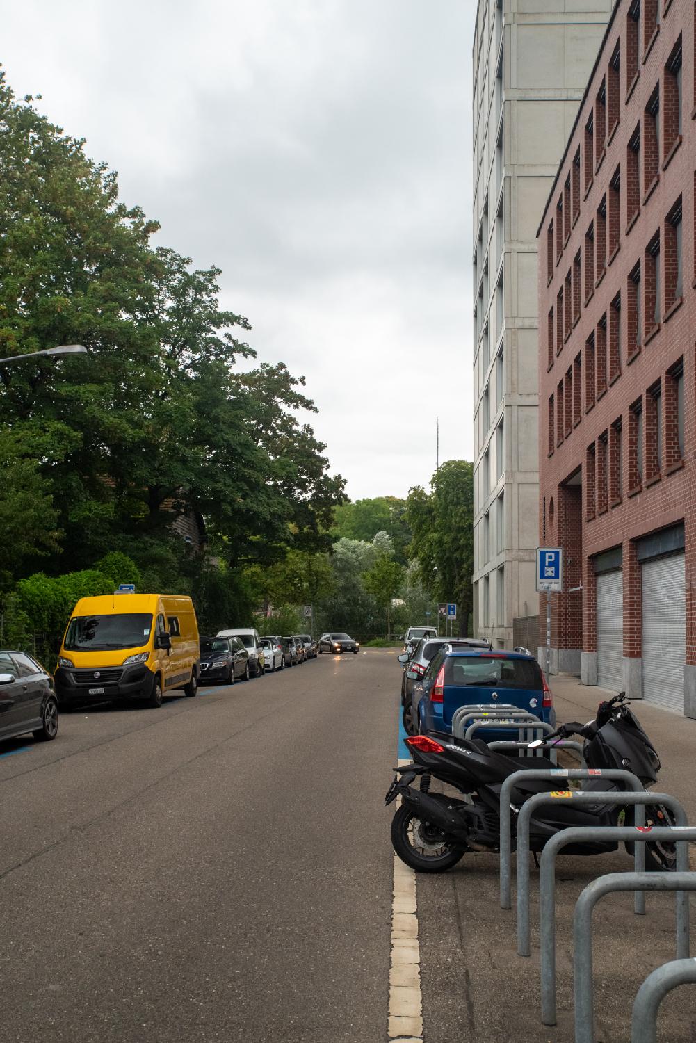 A street with trees and parked cars (including a bright yellow van) on the left, a red and a white building on the right (as well as more parked cars and motorbikes), and a lot of trees in the background after the end of the street. A car is turning on the street towards the photographer.