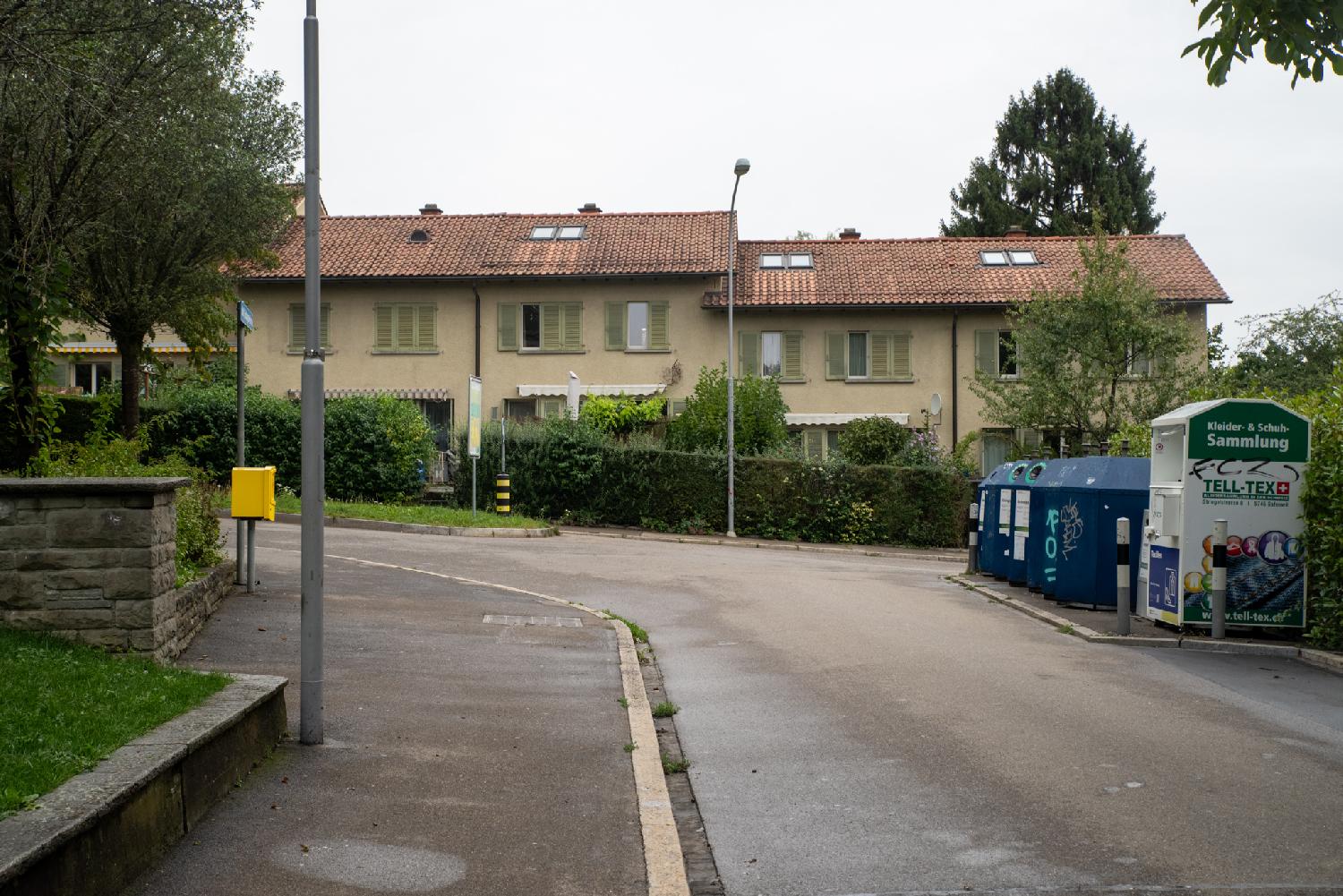 A row of yellow houses with green blinds in a curved street. On the left, there's a yellow mailbox; on the right, recycling containers.