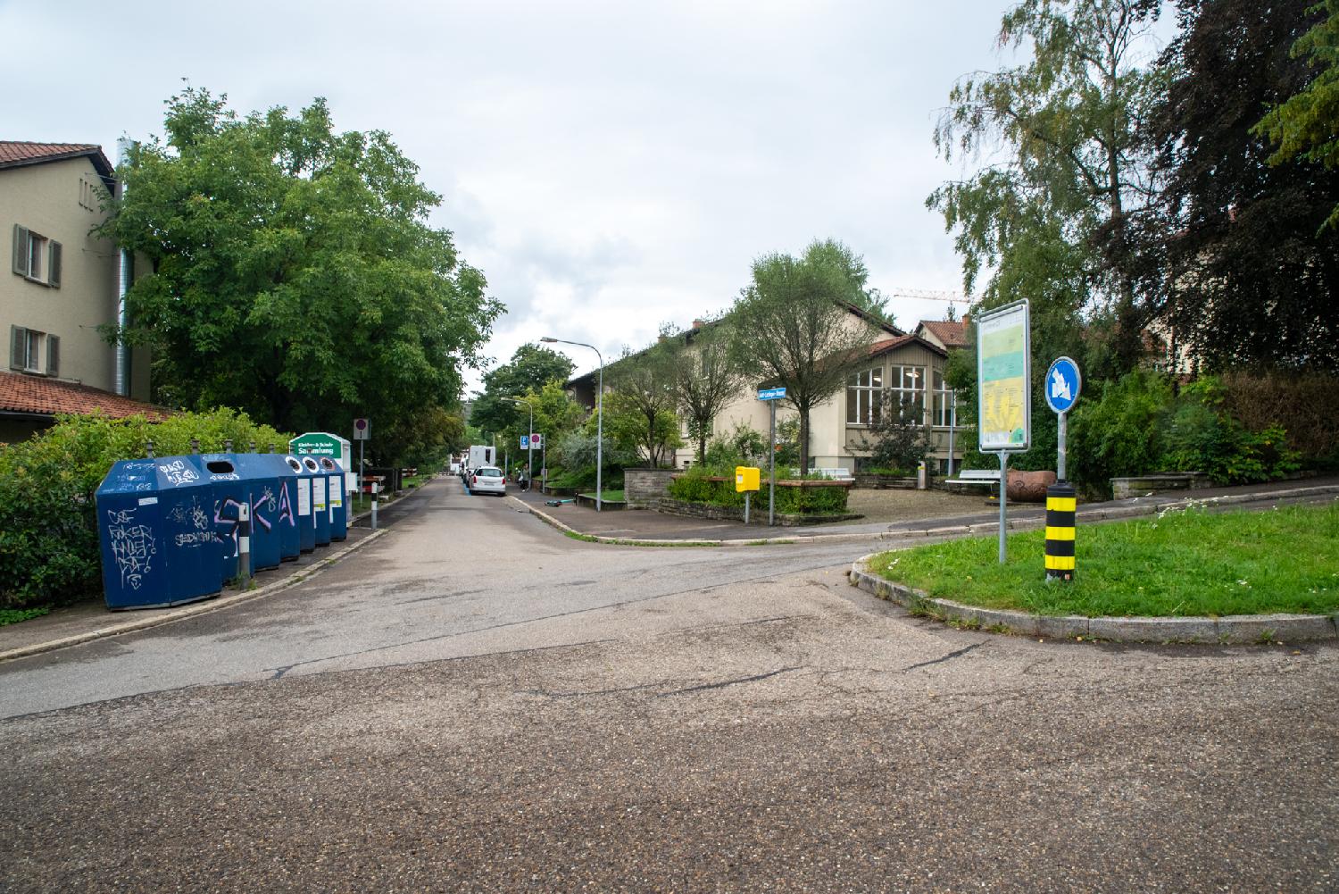 A residential street next to a small square. There are glass recycling bins on the left of the street.