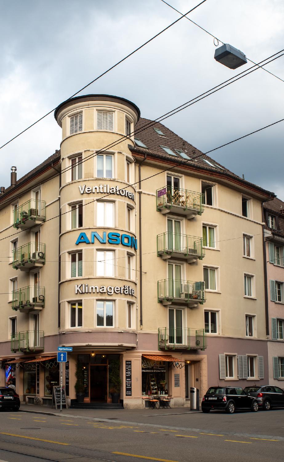 A yellow-beige building with a round column of bay windows forming a tower on the angle of the building. The building has logos for an air conditioning company on the tower and an Italian deli on the ground floor.