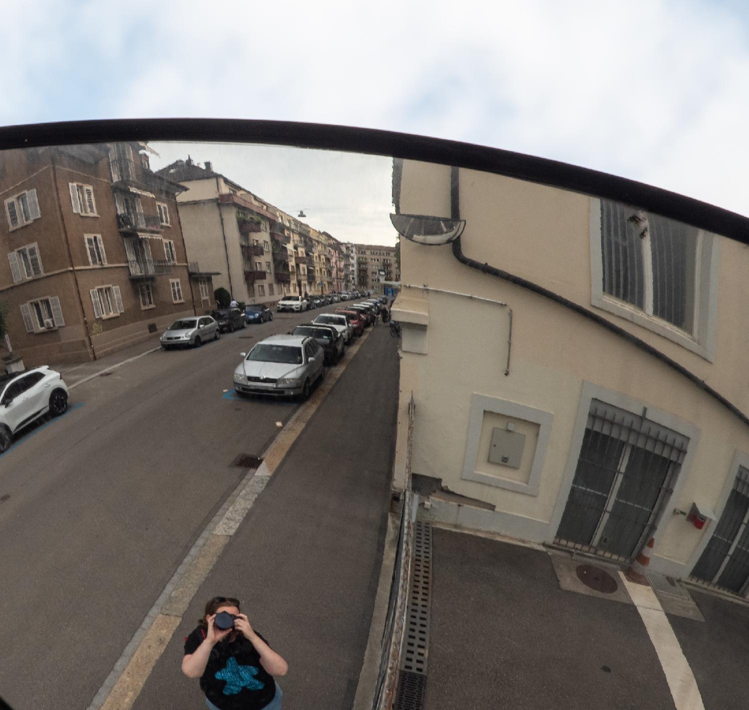 A traffic mirror showing a residential street with 4-stories buildings and cars parked on the street. The photographer is visible behind their camera on the bottom of the picture; the sky is visible above the curved mirror.