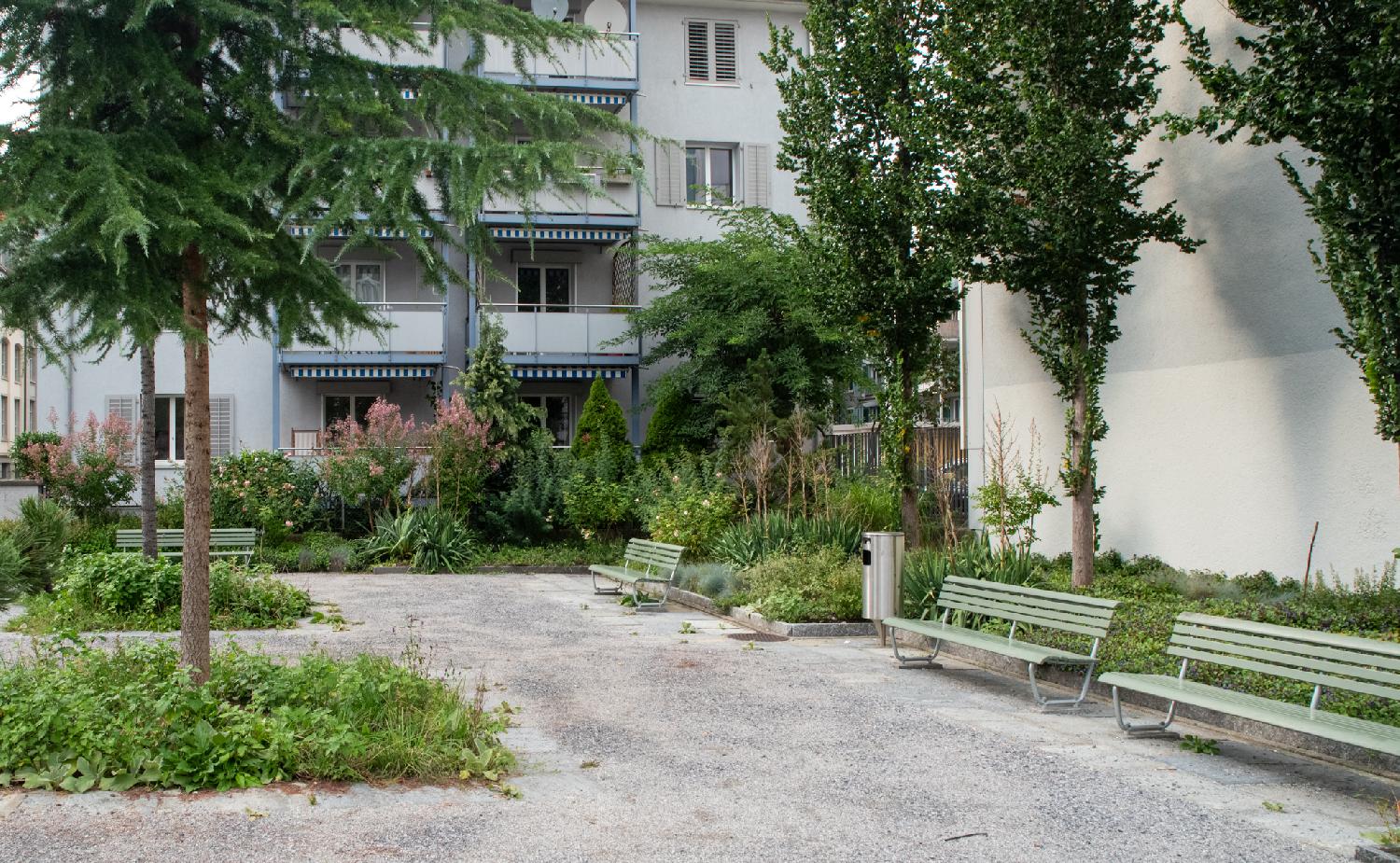 A small park with some greenery, gravel ground and four light-green wooden benches.