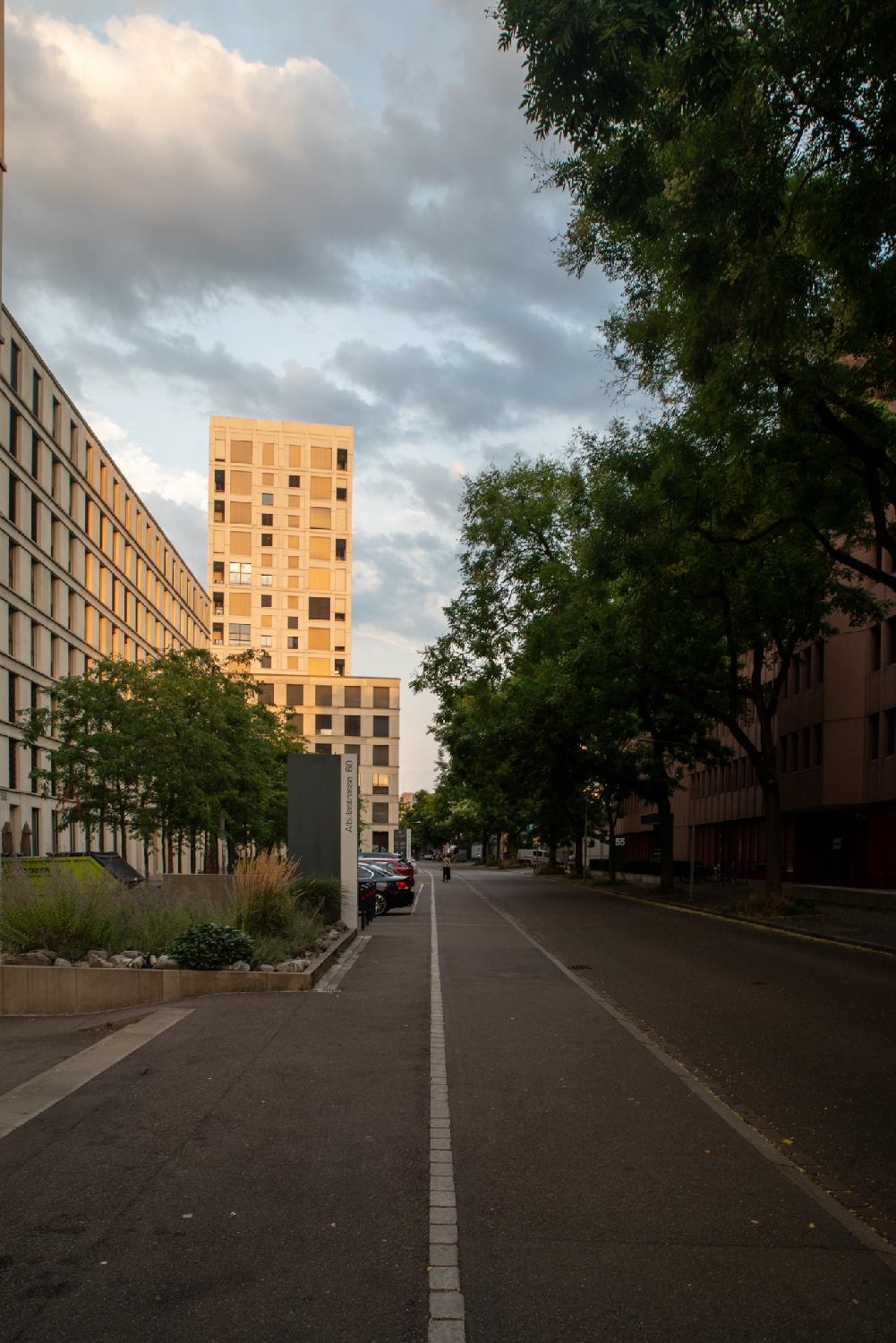 A narrow road with an almost as large sidewalk, with a 8-story building and a small adjoined parking lot on the left, a higher rise building in the background, and trees in front of buildings on the right.