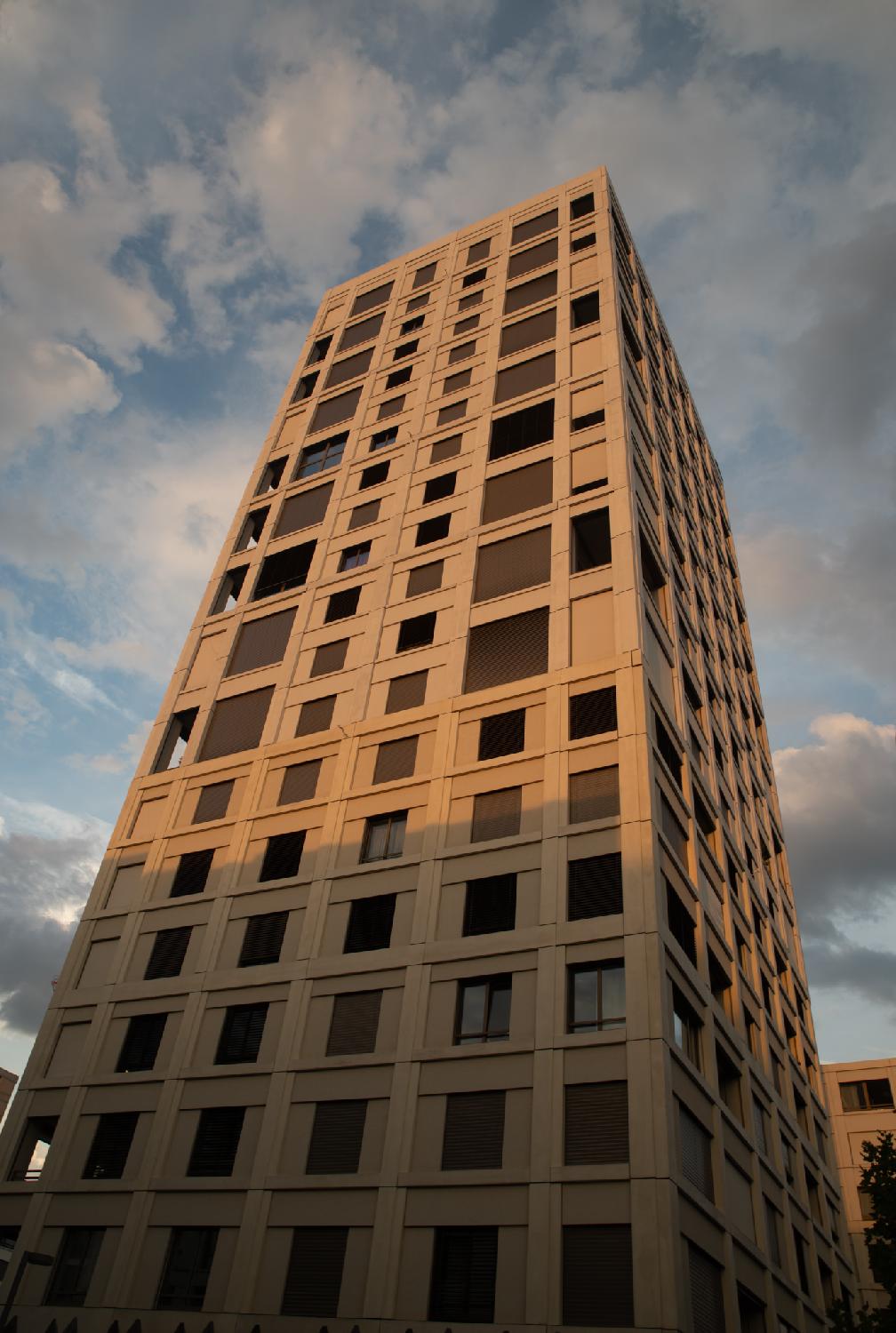 A ~20-story building, seen from the street, with regular columns of windows, most of them with stores down. There's blue, cloudy sky behind the building.
