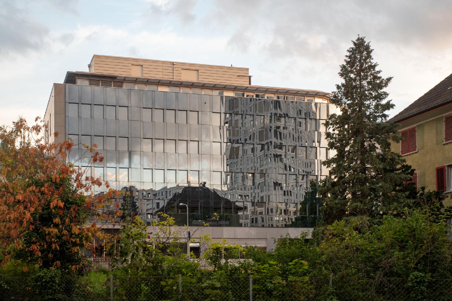 Reflections of a high-rise building on a glass-covered building. There is some greenery in front of the building and a large tree on the right.