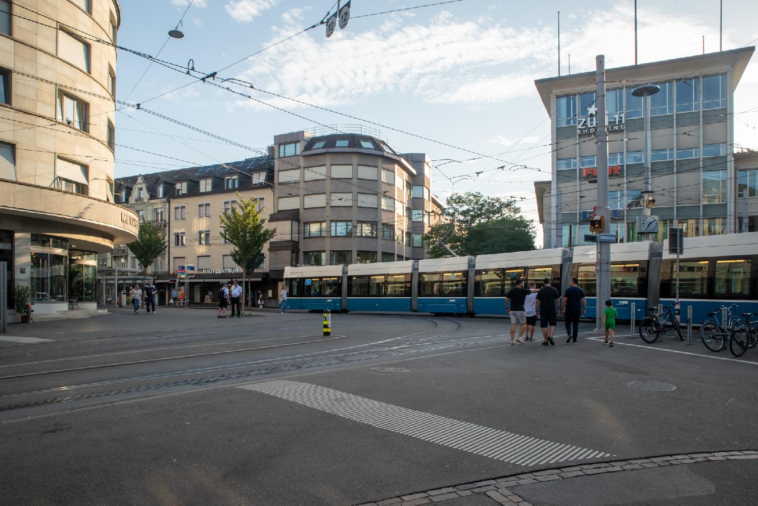 A public square with a few few-stories buildings and a blue Zürich tram arriving at a tram stop.