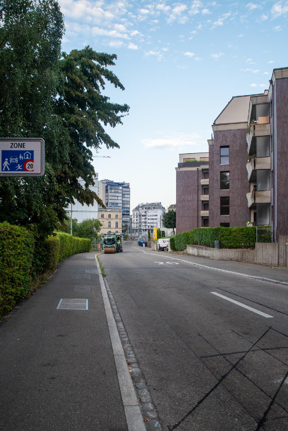 A two way street with a 20km/h limitation painted on the ground and on a sign, hedges on the left, brown-read buildings on the right, and high-rise buildings in the background. There's a light green construction vehicles parked on the left of the street.