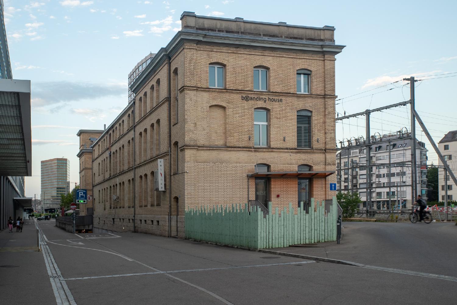 A long, 3-storey yellow/orange brick building on the right side of a street. Train tracks are visible behind it, and a high-rise building appears in the background.