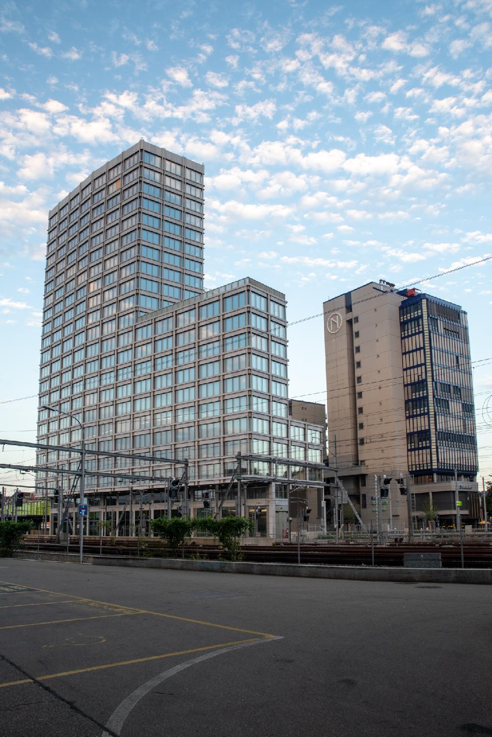 A large L-shaped building with a lot of windows reflecting the blue sky, next to a smaller, older-looking building, above train tracks in Zürich Oerlikon.
