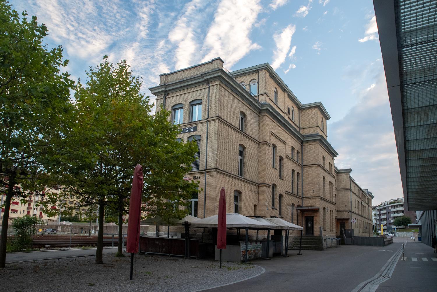 A long, 4-storey yellow/orange brick building on the left side of a street, with trees and a bar terrasse in front of it.