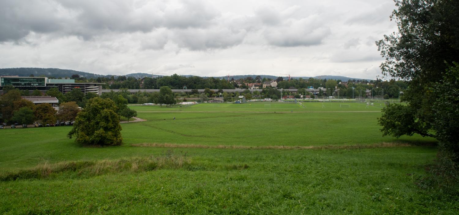 A large green field of grass, with the city in the background. On the left, a mall and its multiple-storey parking lot; a high-raised highway is also visible above a tree line. The further background is hilly.