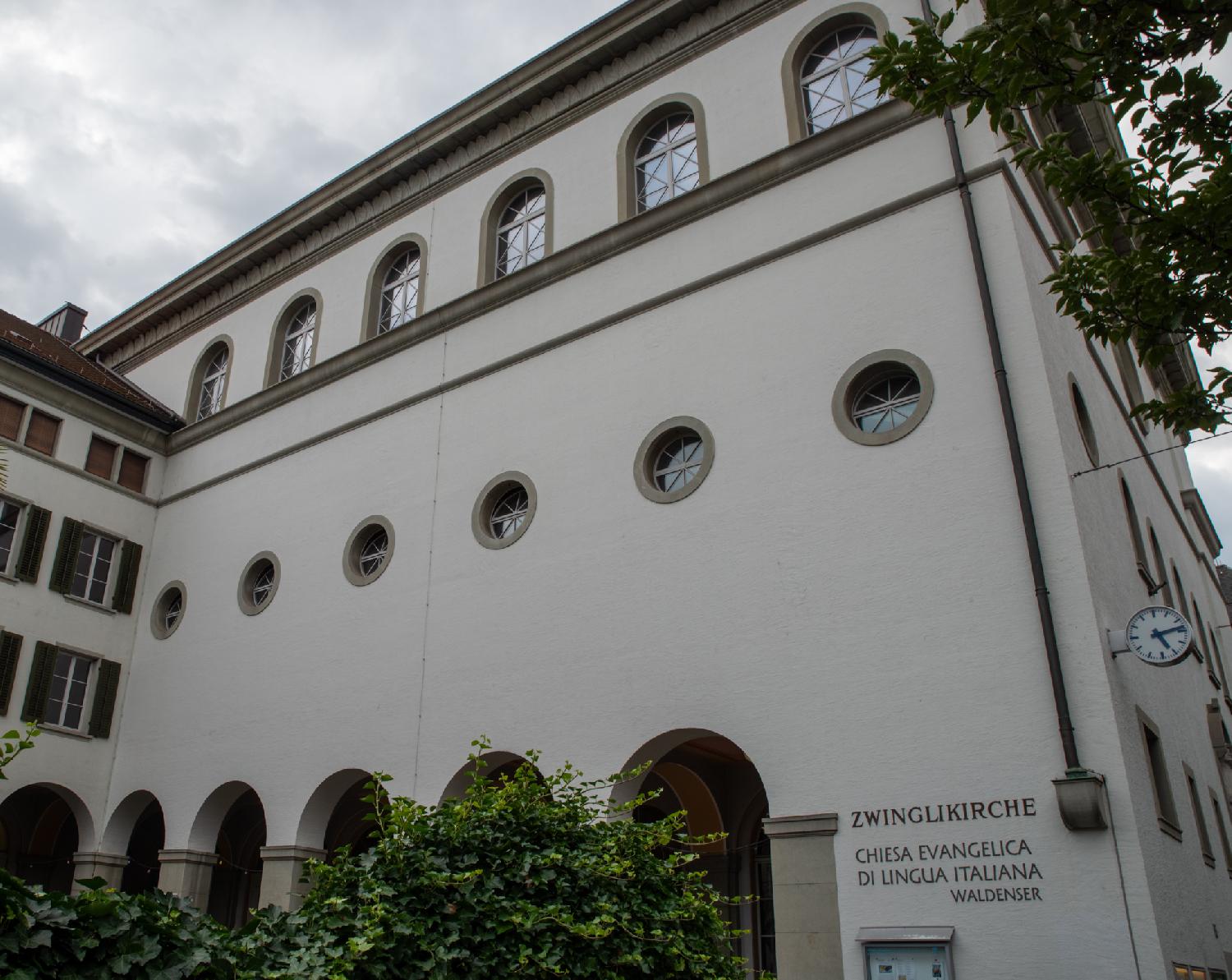 A large white building with arches, round windows and rounded windows above them. An inscription on the building reads "Zwinglikirche - Chiesad Evangelica di Lingua Italiana - Waldenser". A clock on the side of the building shows 5:14.