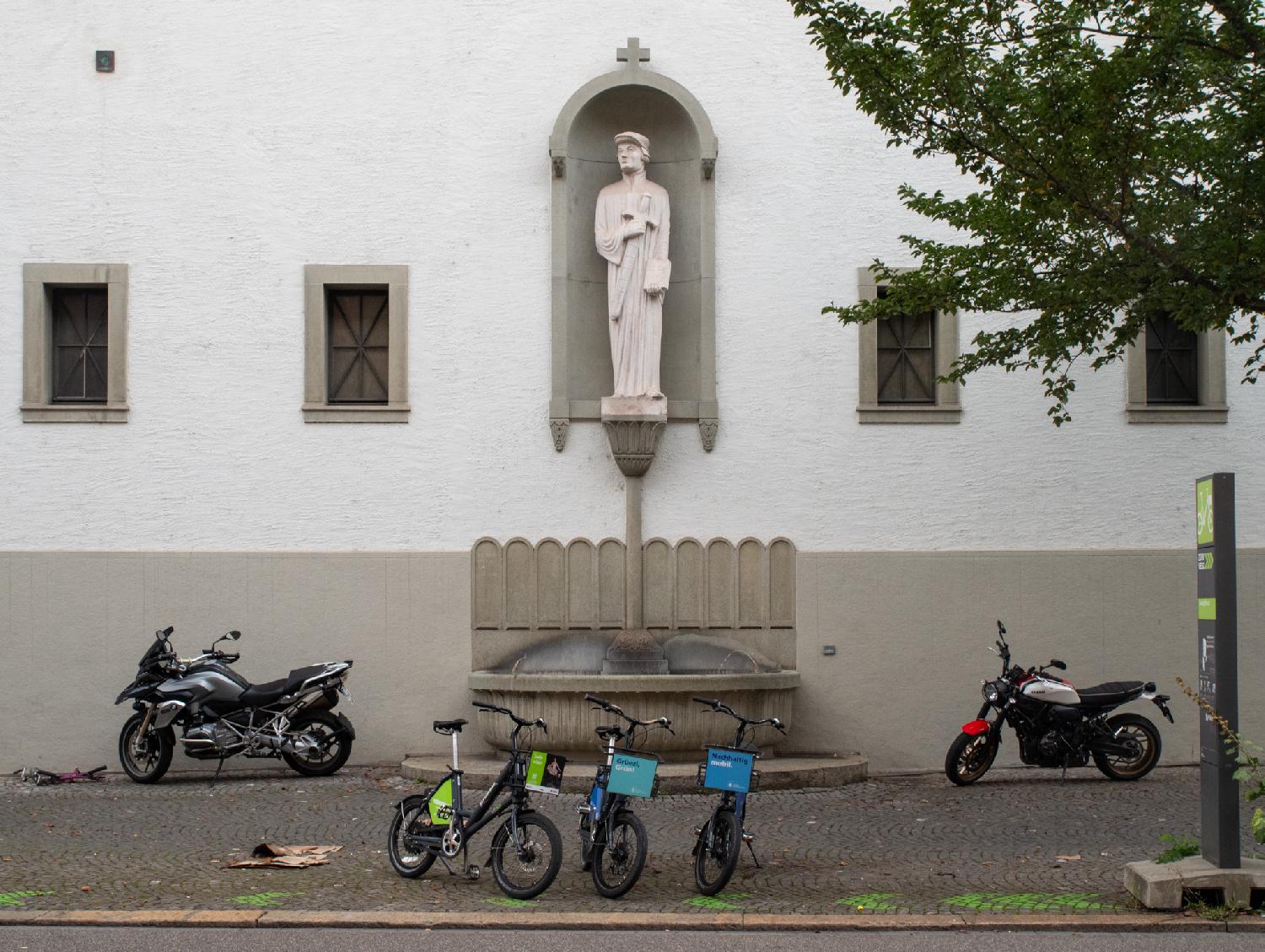 A white statue of a man holding a sword and a book, in an alcove between two windows above a fountain. The picture is taken from the street, where two motorcycles and three city bikes are parked around the fountain.