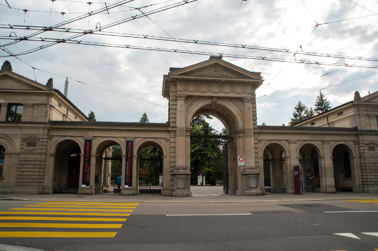A large stone entrance with columns and moulds, composed of a large arch surrounded by three smaller arches on each side. A yellow zebra leads to the left side and trolley cables are visible above the street.