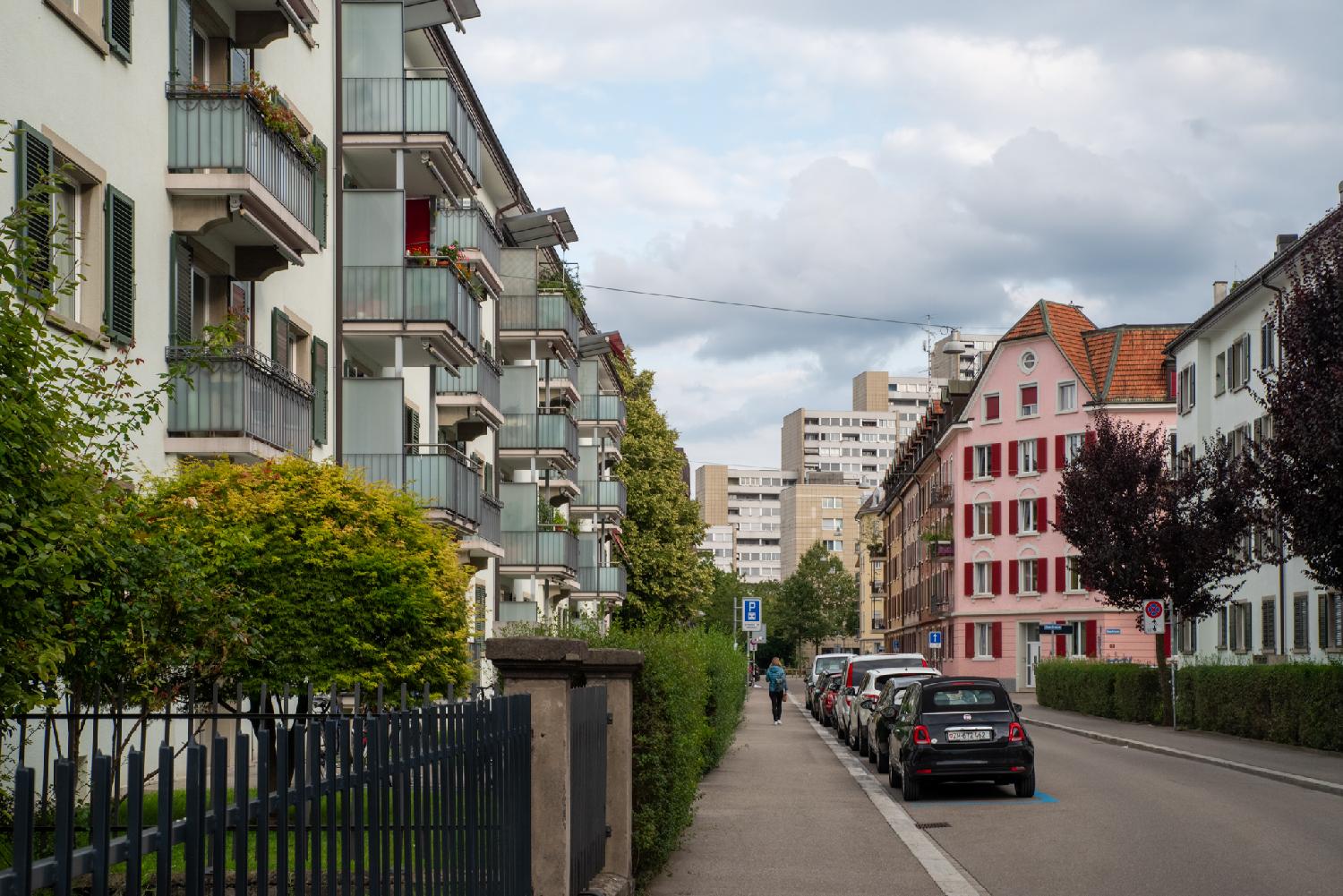 A residential street with 5-storey buildings and cars parked in the street. There's a more visible pink building with red blinds, and the background shows higher-rise buildings.
