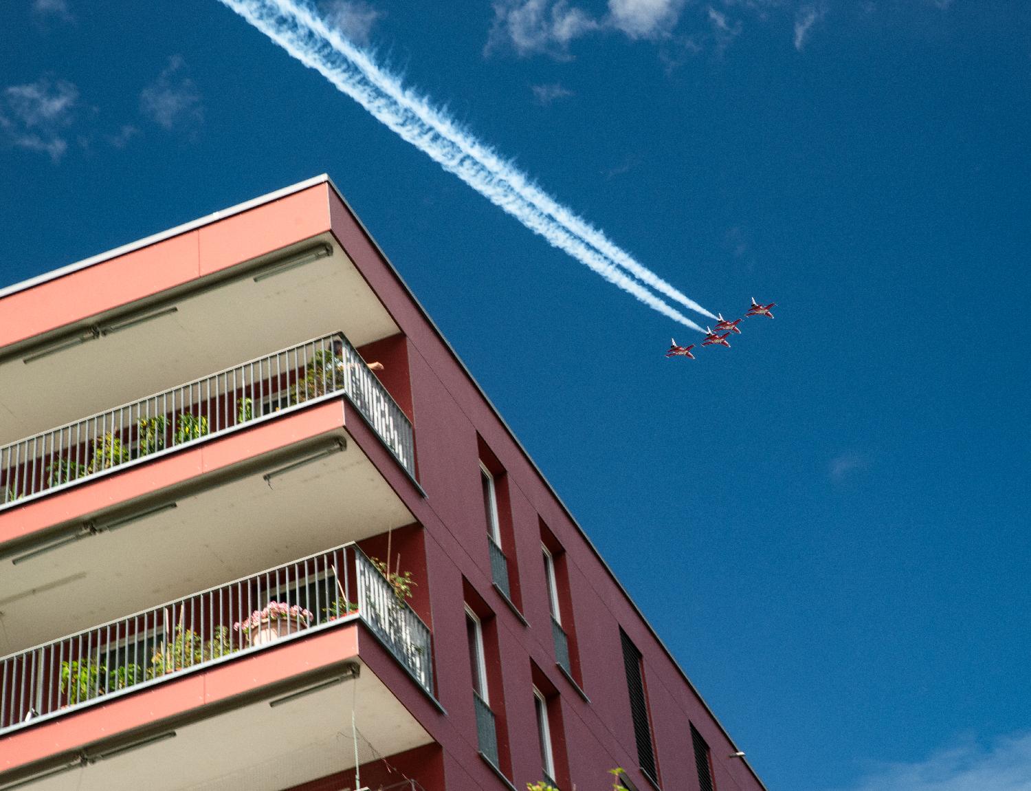 Four red jet airplanes flying in formation in the blue sky above a red building.