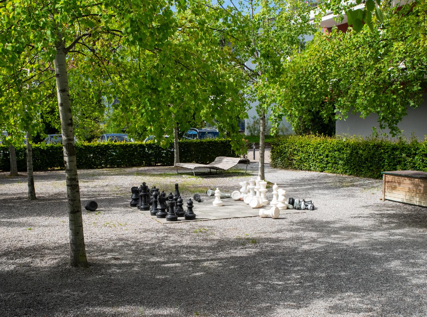 A giant chess set in a park. The pieces are mostly on the board, but some of them have fallen. Trees are providing some shade over the park.