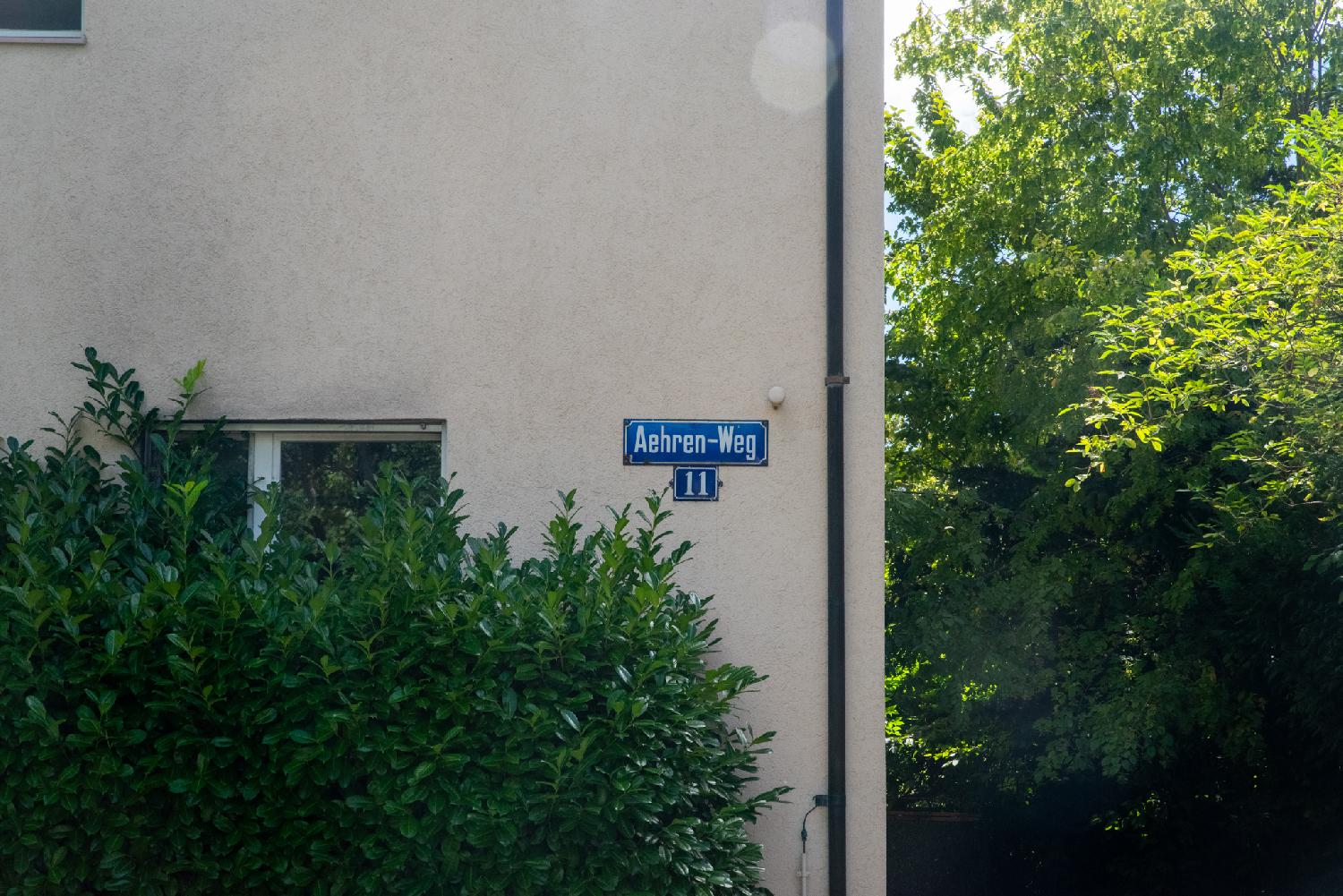 A blue "Aehren-Weg" street sign on a wall, on top of a "11" blue house number plate. The wall is partly hidden by greenery and the right third of the image is filled with trees.