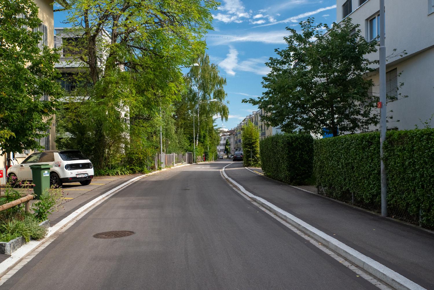 A residential street with 4-storey apartment buildings on each sides, green hedges and trees.