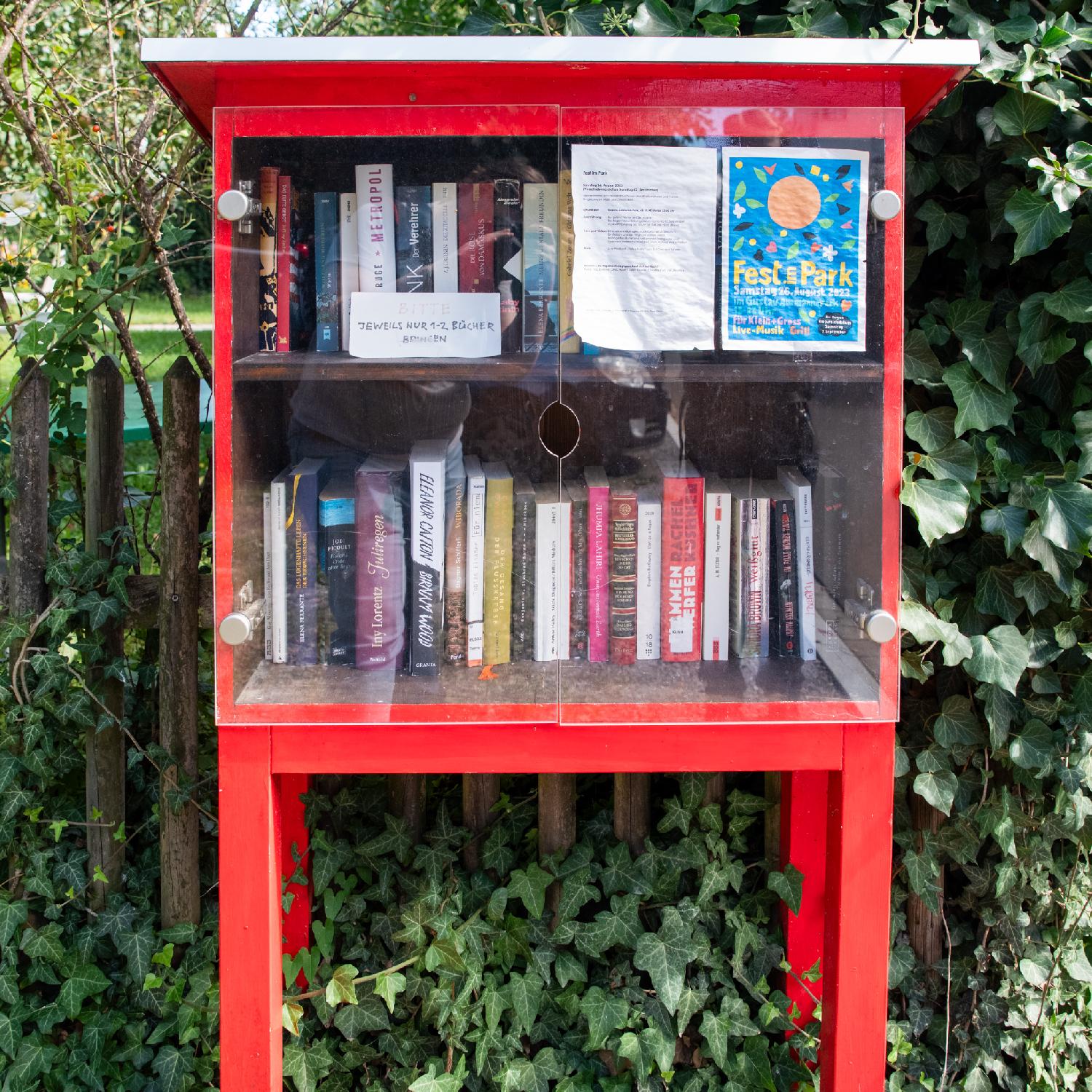 A red bookcase with two shelves full of German-language books behind glass doors. A sign asks "Bitte jeweils nur 1-2 Bücher bringen" - "please only bring 1-2 books at a time". A blue sign for a local festival is also stuck to the glass.