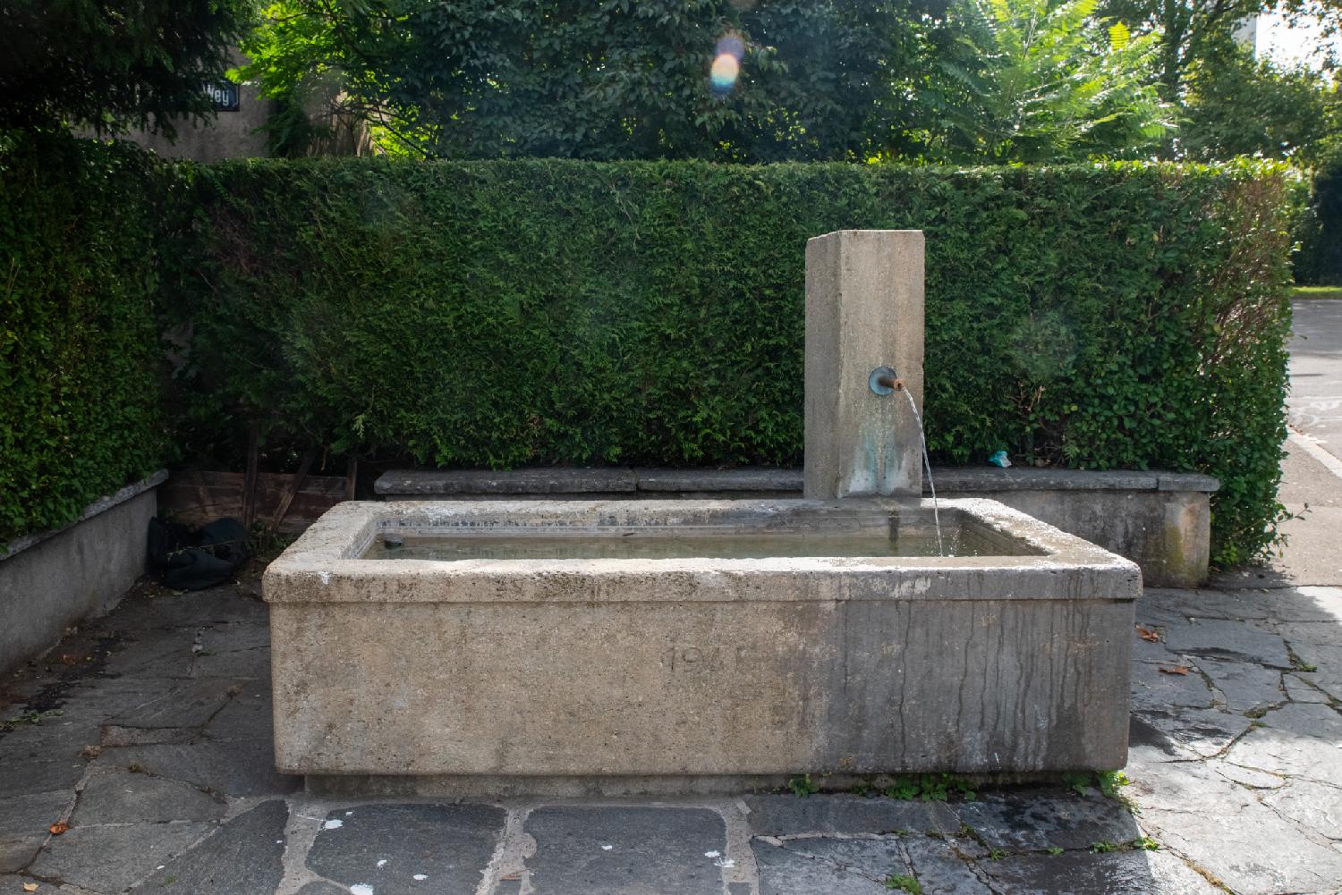 A stone fountain composed of a large rectangular basin and a faucet spewing water from a small pillar on its right. The date 194x is carved on the basin (the last digit is unreadable).
