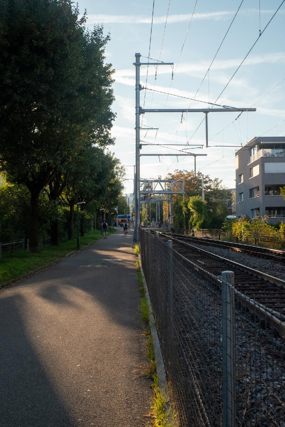 A pedestrian path on the left, separated from a train track on the right by a metallic fence. There are trees on the left of the path, and a grey building on the right of the train tracks.