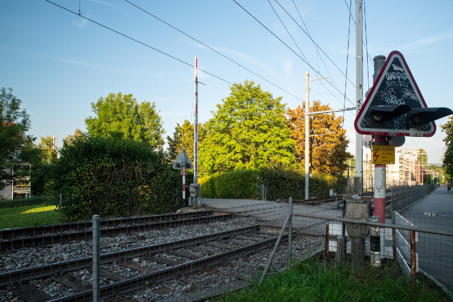 Two train tracks crossed perpendicularly by a road. There are open barriers on each side of the tracks and a light signal indicating when a train is incoming.
