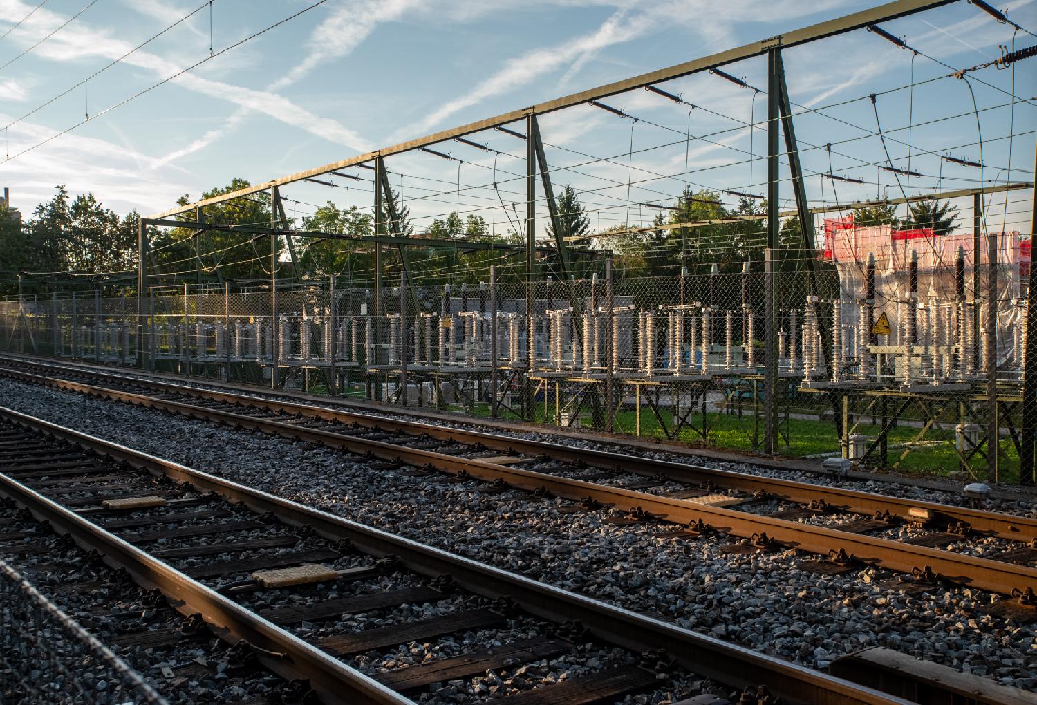 A power installation made of a lot of cables and large coils, behind a fence, behind rail tracks.