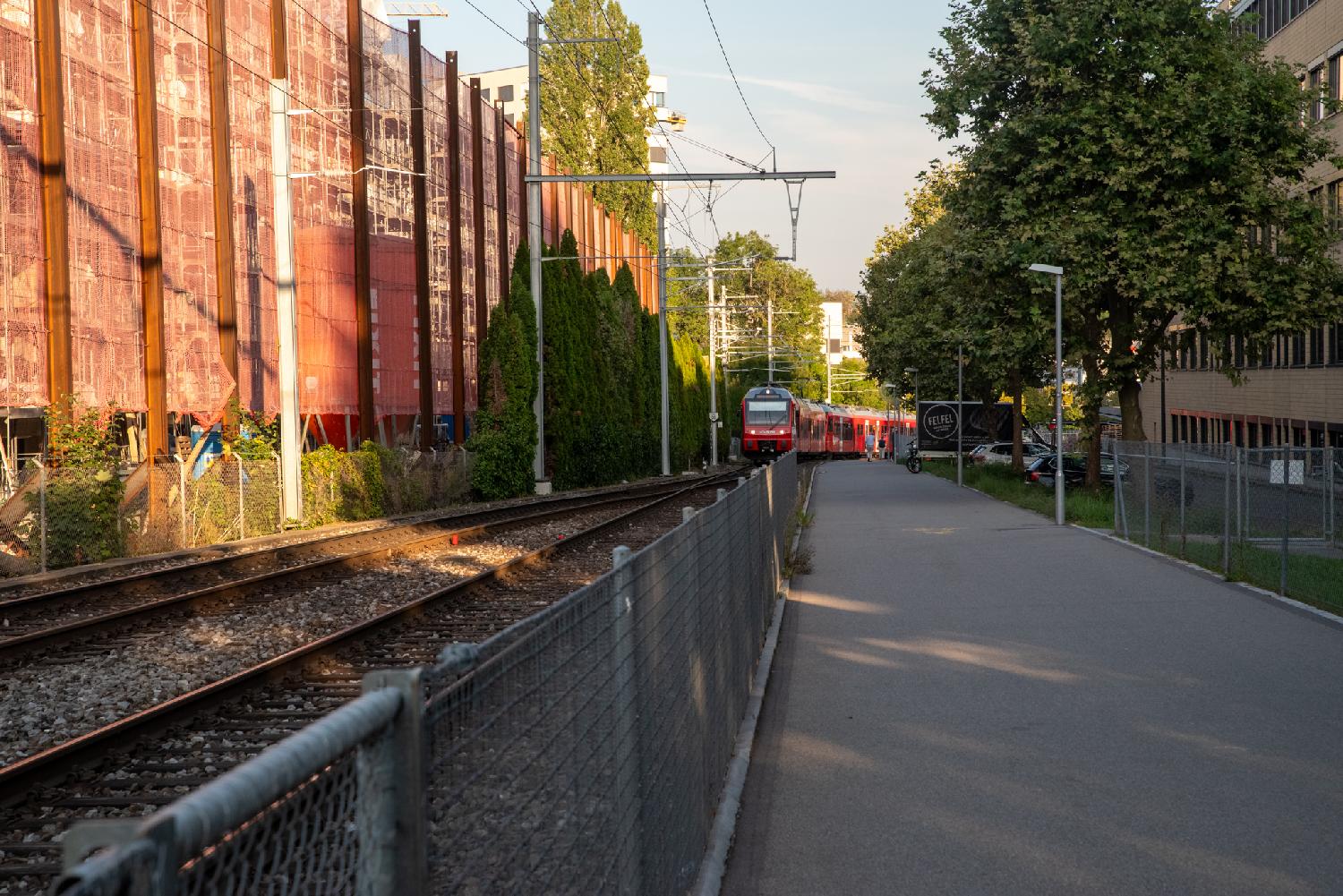 A pedestrian path on the right, a fence and rail tracks, on which a red train is incoming in the background. There's an orange works fence on the left of the train track.
