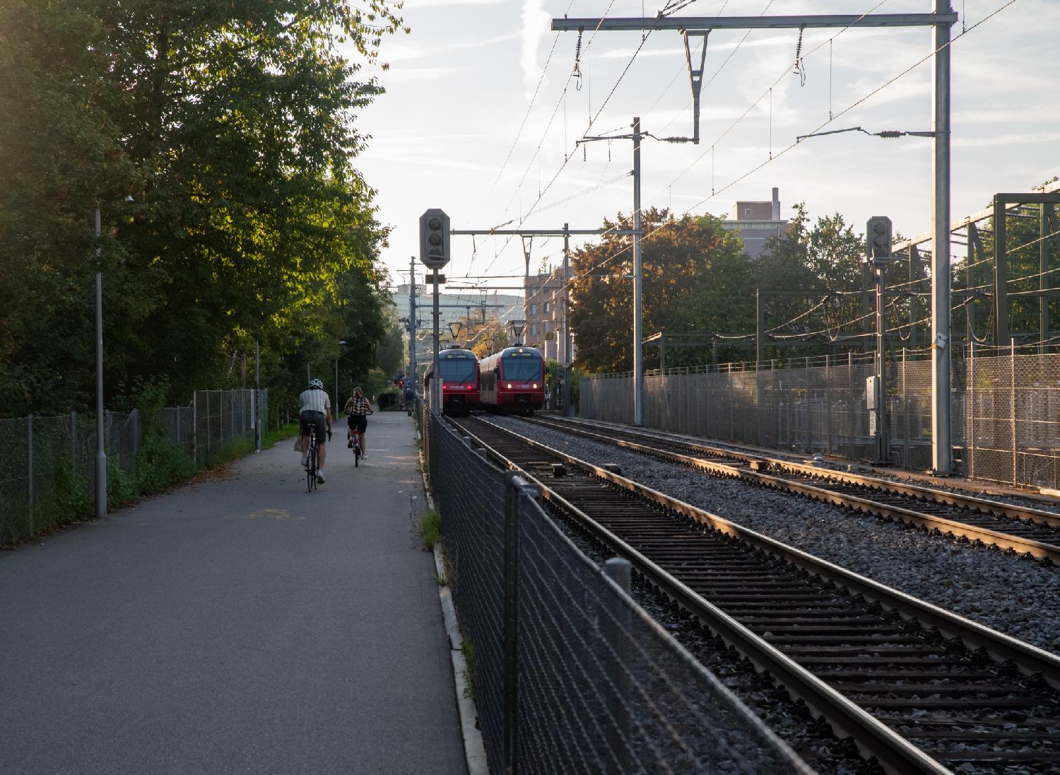 A pedestrian path with a couple of bikes on it on the right, separated from two train tracks by a metallic fence. There are two red trains on the tracks, in opposite directions: one goes to Uetliberg, the other to Zürich HB.