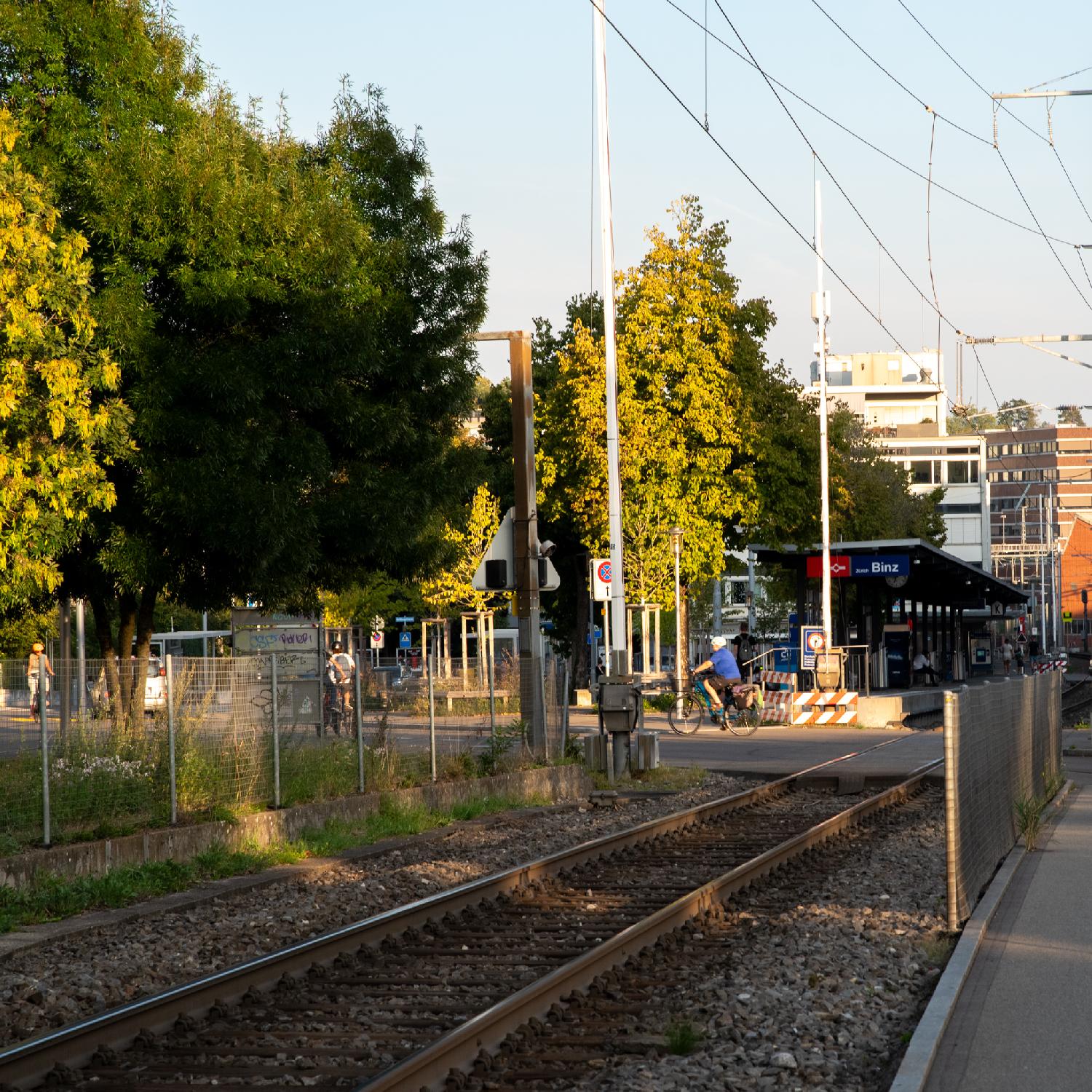 A train track, a level crossing with barriers, and the Zürich Binz train station (a single platform along the track) in the background.