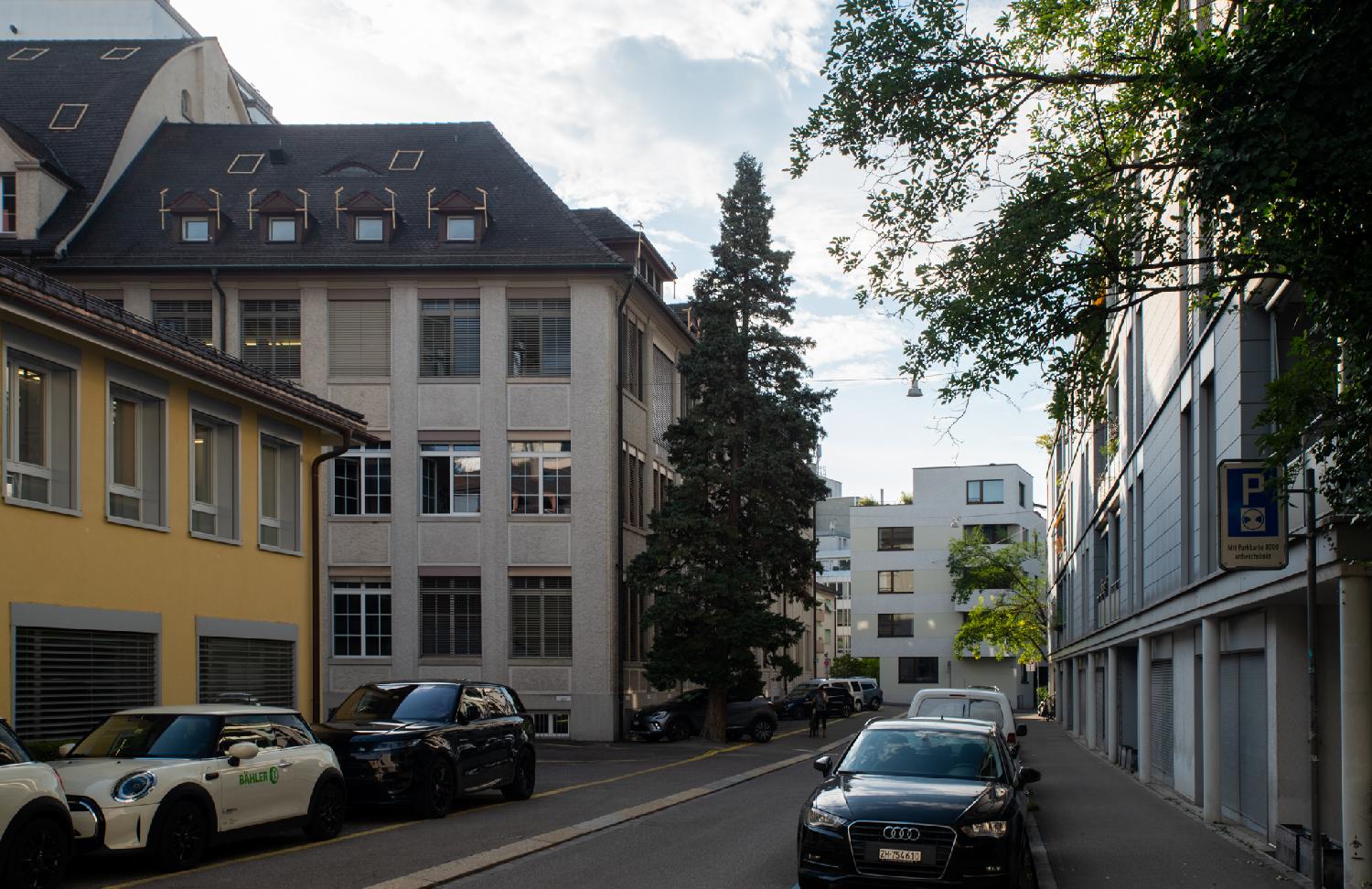 A narrow street with 4-storey buildings and cars on each side, and a 3-4-storey-building in the background.