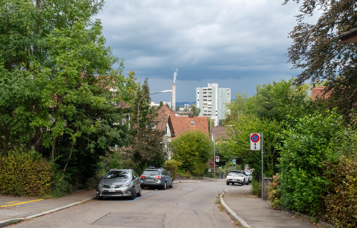A curved road, with cars parked on blue parking spots on both sides of the road. There's a high rise and a smoking industrial chimney in the background, and the sky is threatening.