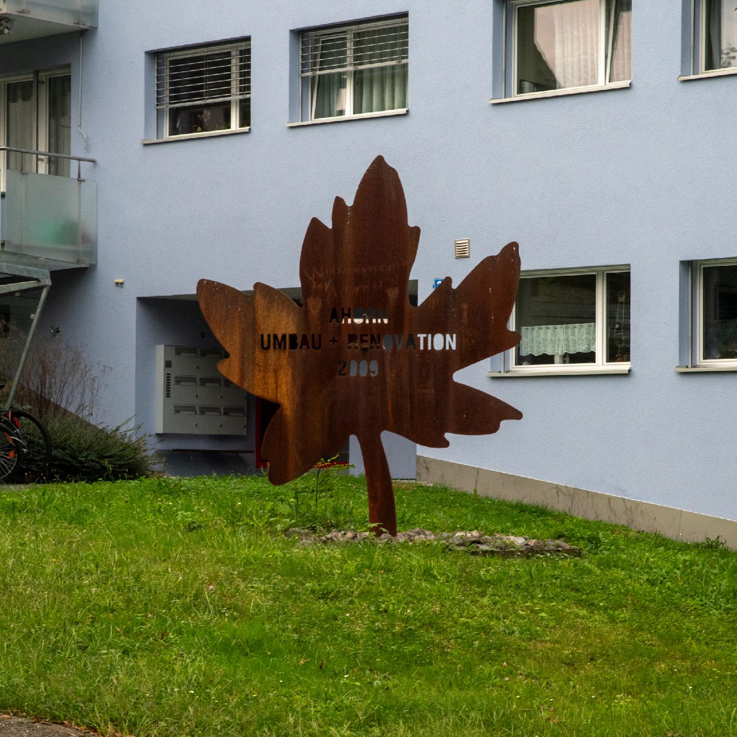 In front of a blue-grey building, in the lawn, a large rusty metallic pane representing a maple tree, with the words Ahorn - Umbau + Renovation - 2009.