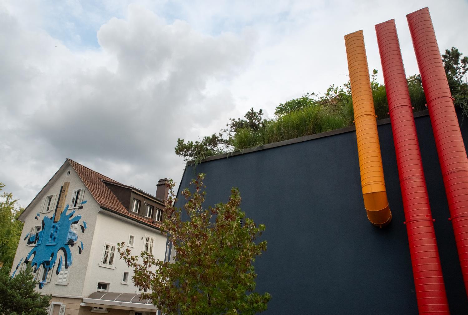 Two buildings - one is a white house with painting on a facade (a large blue splotch held by a wooden laundry pin), the other one is a more industrial-looking building with three pipes (one orange and two red) stuck on the facade.