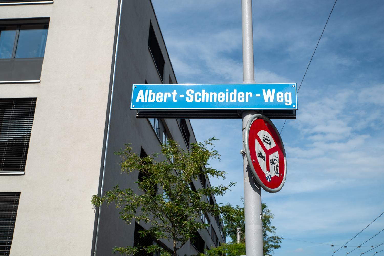 A blue street sign on a pole, indicating Albert-Schneider-Weg. The pole also hosts a sign indicating that no traffic is allowed on the street. The background has a grey building and blue sky.