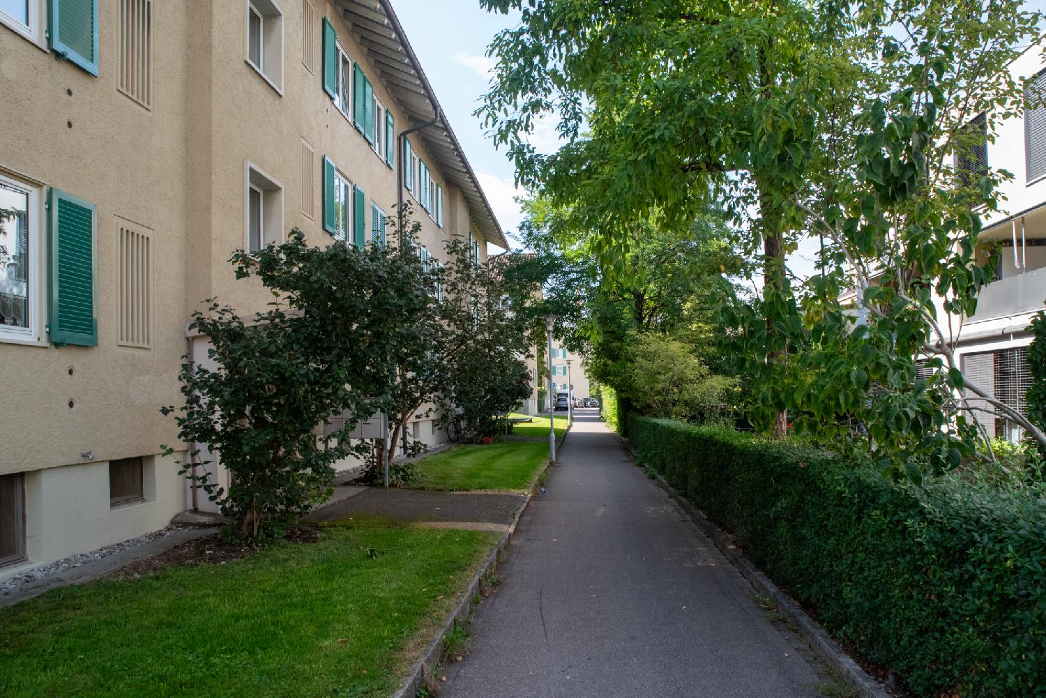 A pedestrian alley with a 3-storey yellow building with green blinds on the left, a green hedge on the right, and a white building behind the hedge.